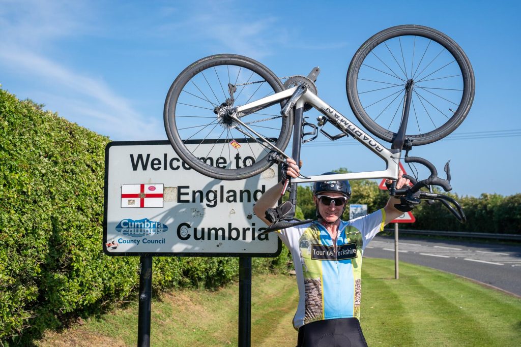 A participant lifting a bike above the Welcome to England sign